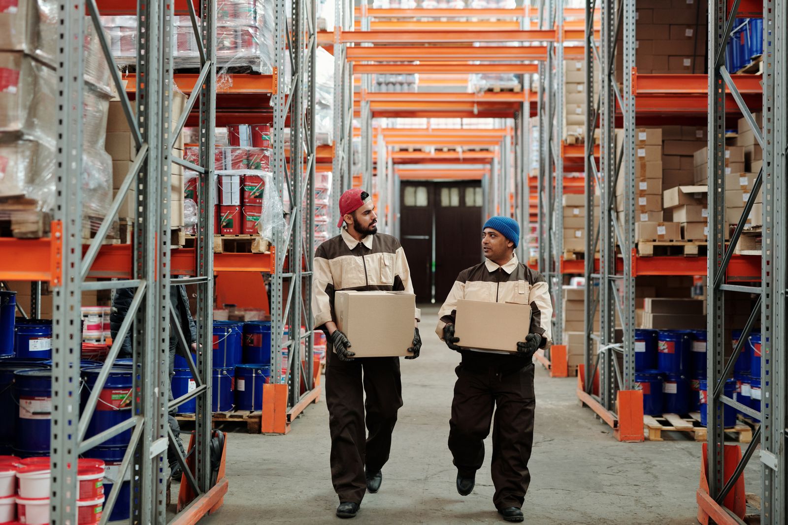 Two warehouse workers carrying cartons through a storage aisle